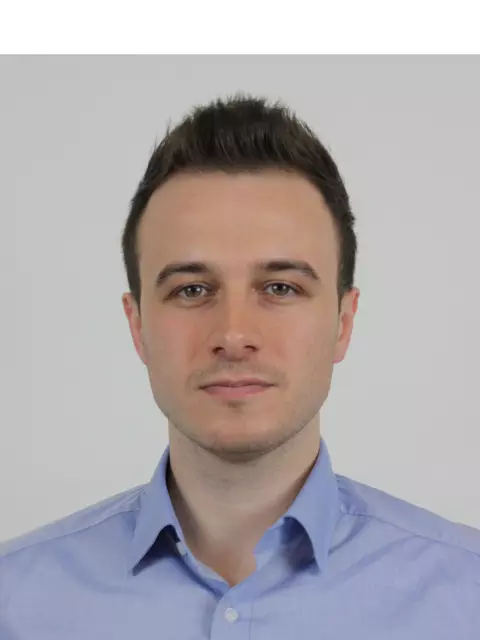 Ein Mann mit dunklen kurzen Haaren steht vor einer weißen Wand.